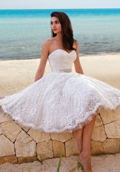 a woman in a short white dress sitting on a stone wall near the ocean with her legs crossed