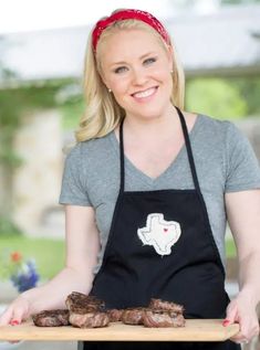 a woman in an apron is holding some meat on a cutting board and smiling at the camera