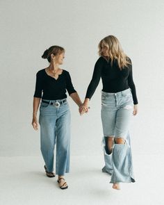 two women are holding hands and walking towards the camera, both wearing high waisted jeans