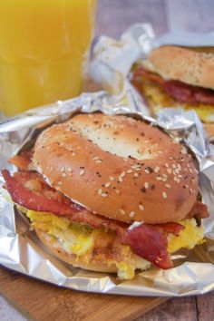 two sandwiches with bacon, eggs and cheese are on aluminum foil next to a glass of orange juice