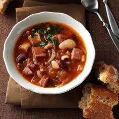 a bowl of soup with meat, beans and bread next to it on a napkin