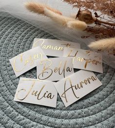 four white and gold name tags sitting on top of a gray blanket next to dried plants