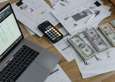 an open laptop computer sitting on top of a wooden desk covered in bills and money