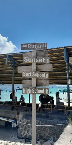 the sign is pointing in different directions at the beachside area with people sitting and standing around