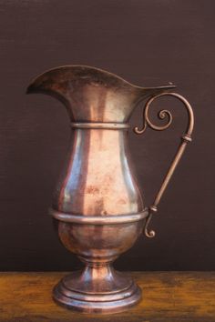 an antique silver pitcher sitting on top of a wooden table next to a brown wall