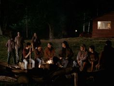 a group of people sitting around a fire pit in the woods at night with lights on