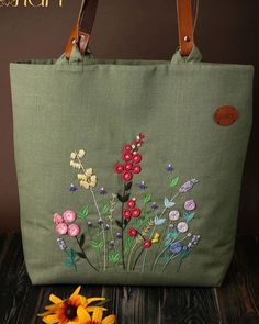 a green bag with flowers embroidered on the side and leather handles, sitting next to a yellow flower