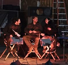 three people sitting on folding chairs in front of a stage with microphones around them