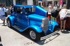 an old blue car parked in front of a building with people standing around and looking at it