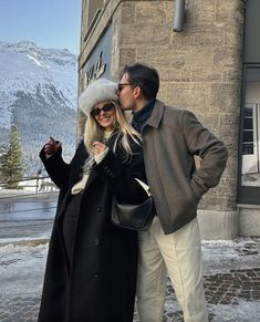 a man and woman standing next to each other in front of a building with snow on the ground