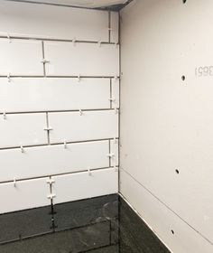 the corner of a white tiled room with black flooring and exposed pipes on the wall