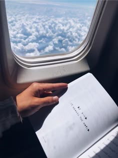 a person holding an open book in their hand while looking out the window above the clouds