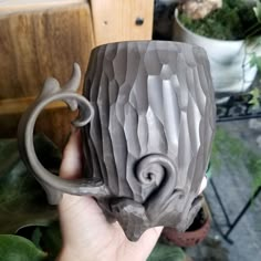 a hand holding a gray ceramic mug in front of a potted plant