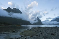 the mountains are covered in clouds and low lying rocks at the edge of a body of water