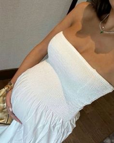 a woman wearing a white dress and holding a gold purse in her right hand while sitting on a chair