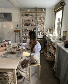 a woman sitting at a table in a room with pottery on the walls and shelves