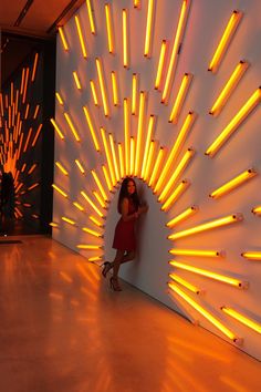 a woman standing in front of a wall with yellow lights on it and her arms behind her back