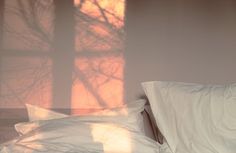 a bed with white sheets and pillows in front of a window that has the sun shining through it