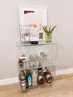 a clear shelf filled with bottles and glasses on top of a hard wood floor next to a white wall