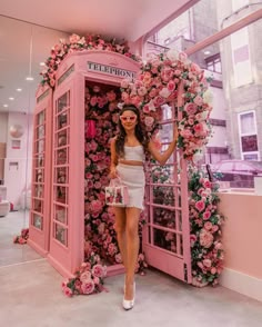 a woman standing in front of a phone booth with pink flowers on the wall behind her