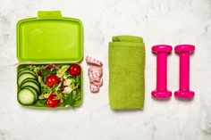 a green lunch box filled with vegetables and fruit next to two pink utensils