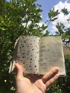 an open notebook with writing on it in someone's hand near some trees and bushes