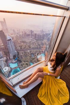 a woman in a yellow dress sitting on the edge of a window looking out over a city