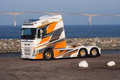 an orange and white semi truck parked next to the ocean with a bridge in the background