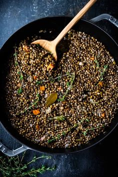 a pan filled with lentils and herbs on top of a black surface next to a wooden spoon