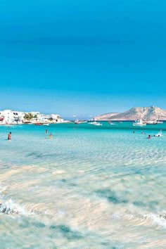 people are swimming in the clear blue water on a beach with boats and mountains in the background