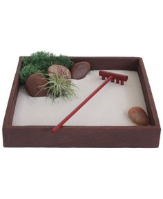 a wooden tray with rocks, plants and a rake in it that is filled with sand