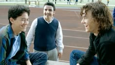 three young men sitting on the side of a tennis court talking to each other and laughing