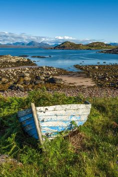 an old boat is sitting on the grass by the water's edge, with mountains in the background