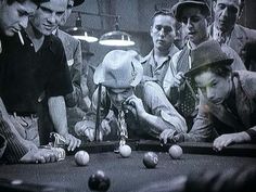 a group of men standing around a pool table with balls on it and one man wearing a hat