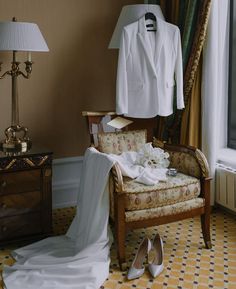 a wedding dress and suit hanging on a hanger next to a chair in front of a window