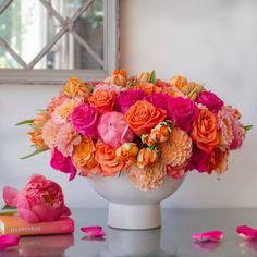 a white vase filled with pink and orange flowers on top of a table next to a book