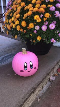 a pink pumpkin sitting on the sidewalk next to some yellow and purple flowers in front of a potted plant