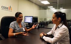 two women sitting at a table talking to each other in an office setting with computers on the wall