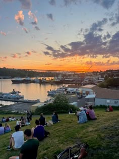 many people are sitting on the grass by the water at sunset with boats in the background