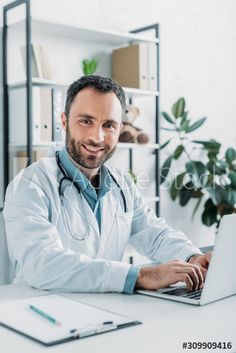 a man in a lab coat is sitting at a desk with a laptop and smiling