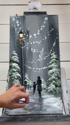 a person is painting a scene on a canvas with snow and christmas trees in the background