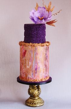 a three tiered cake with purple icing and flowers on top, sitting on a gold pedestal