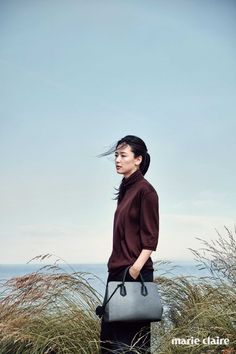 a woman standing in tall grass holding a gray handbag and looking off into the distance