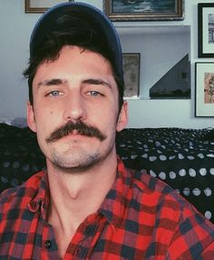 a man with a moustache wearing a hat and looking at the camera while sitting on a couch