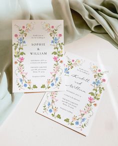 two wedding cards with flowers on them sitting on top of a white cloth covered table