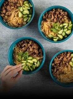 four bowls filled with noodles and vegetables on top of a gray countertop next to a person's hand