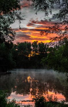 the sun is setting over a lake with trees around it and clouds in the sky