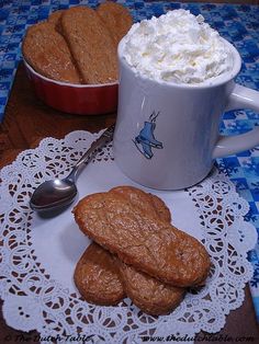 two mugs with whipped cream and cookies on a doily next to one another