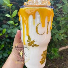 a hand holding up a glass with yellow liquid and honey on the bottom, in front of some bushes