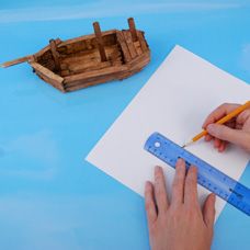 a person is drawing on paper with a pencil and ruler next to a wooden boat